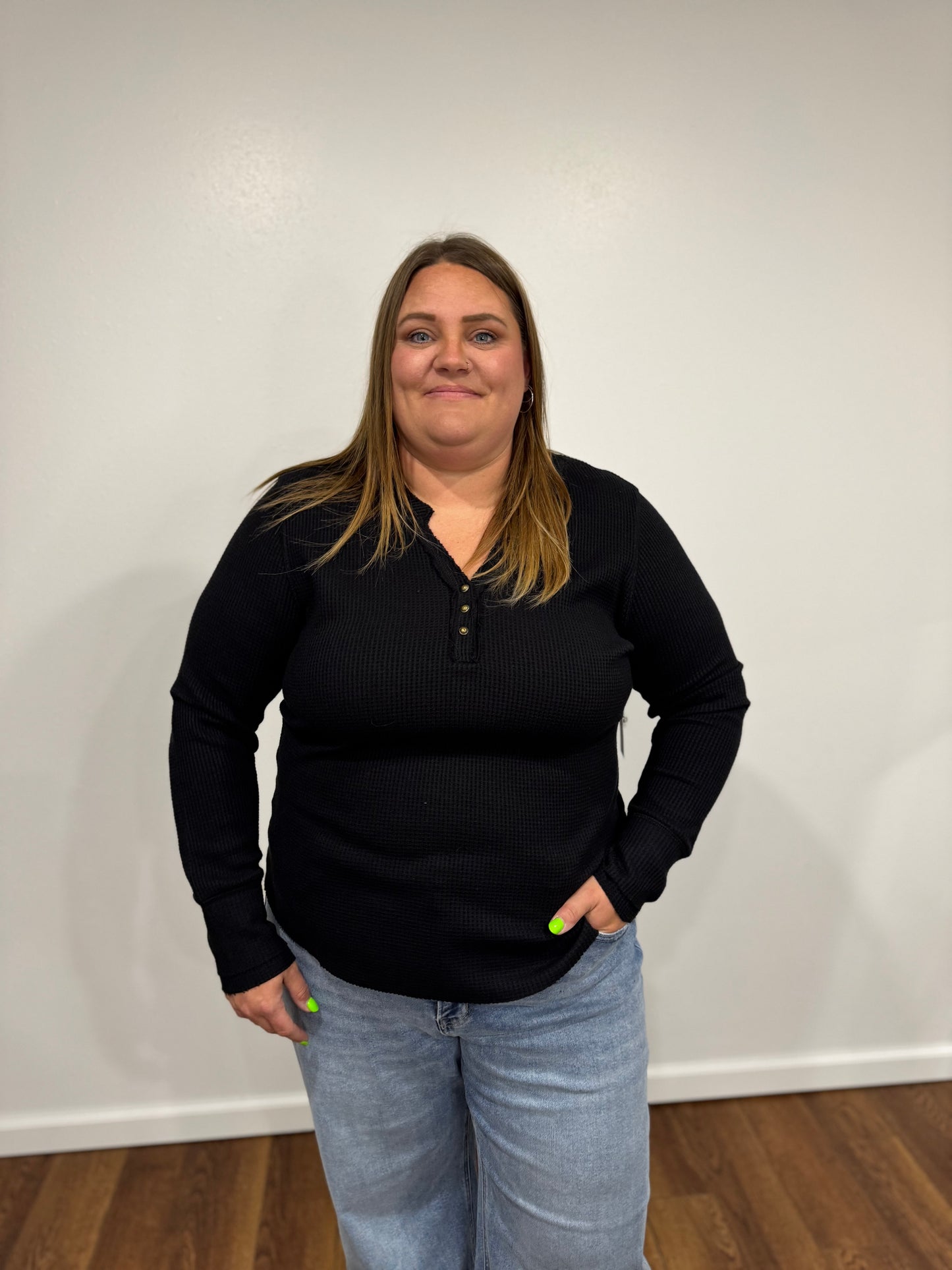 a black long-sleeve henley shirt and blue jeans standing against a white wall.
