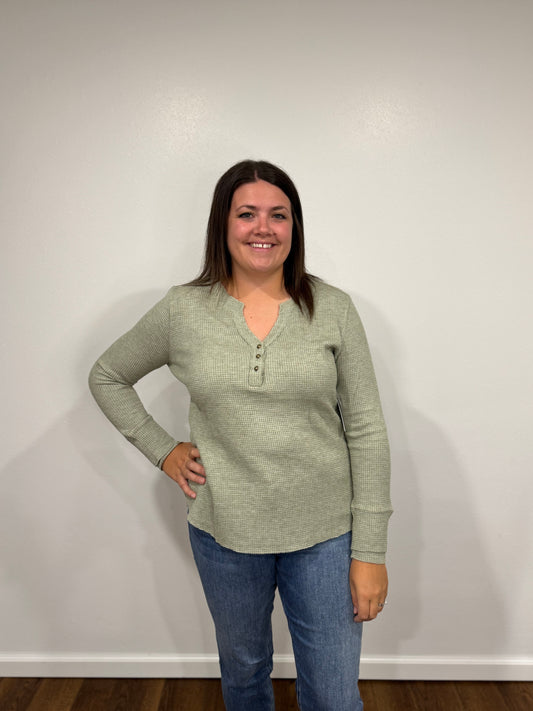 Woman wearing a green long-sleeve henley shirt and blue jeans against a white wall.
