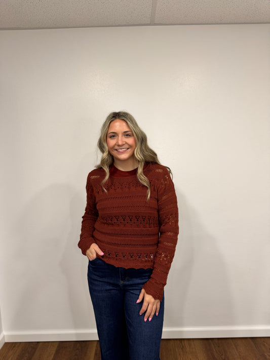 Woman wearing a brown burnt orange sweater and dark jeans standing against a white wall.