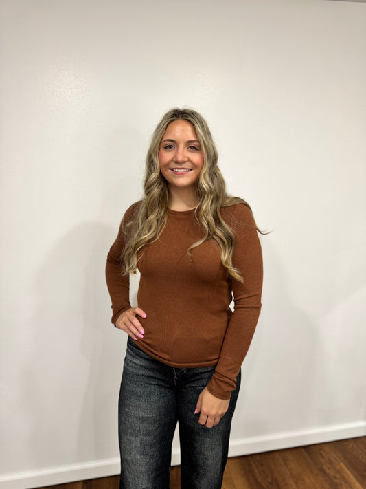Woman wearing a brown sweater and jeans standing against a white wall.