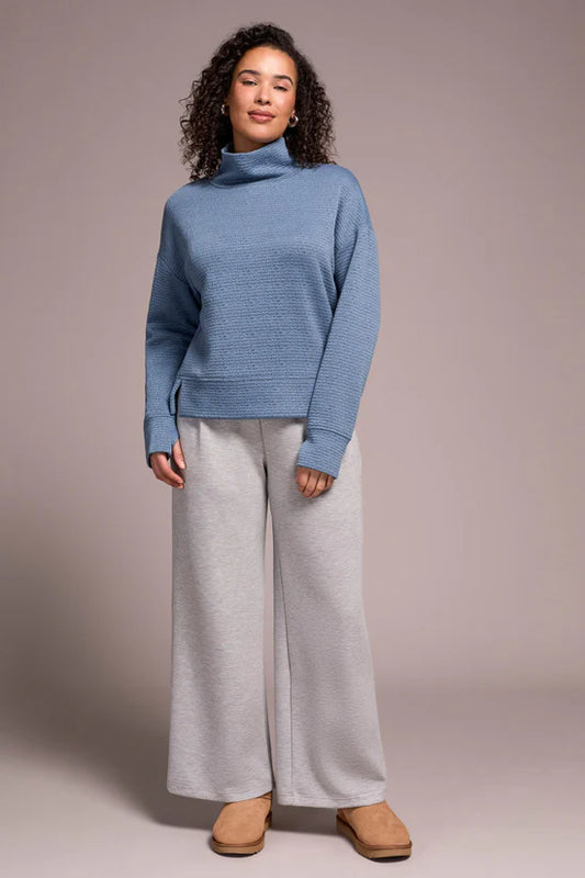Woman wearing a blue sweater and gray pants against a plain background